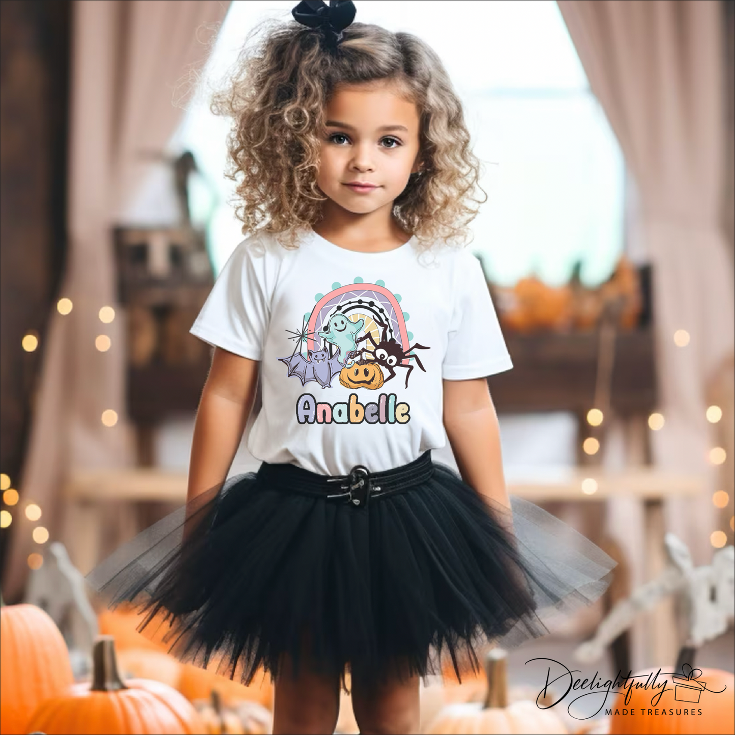 Girl wearing a Bella + Canvas white shirt with a Halloween design that has a bat, ghost, pumpkin, and spider on a rainbow with name.
